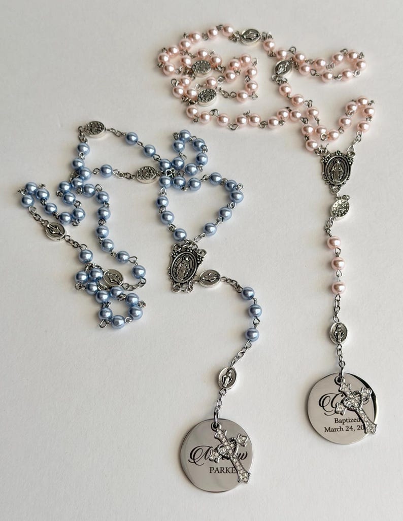 May include: Two rosaries featuring light blue and light pink pearl-like beads on silver-toned metal chains. Each rosary includes a central religious medal and a circular silver pendant engraved with text and a small rhinestone-encrusted cross charm.