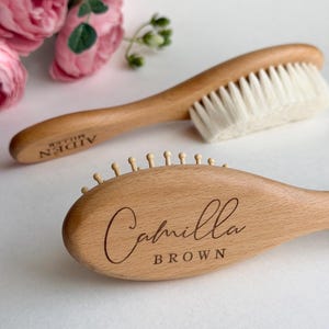 May include: Two wooden baby brushes with light-colored bristles and wooden handles. One brush is engraved with the name "Camilla Brown", and the other with "AIDEN MILLER". Pink flowers are in the background.