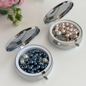 May include: Two round, silver-toned metal cases with hinged lids and interior mirrors. One case contains a rosary with dark blue and white beads, while the other holds a rosary with light pink and white beads, each featuring a small metal crucifix.