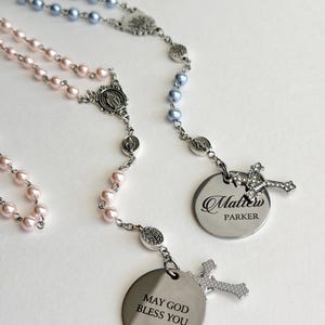 May include: Two rosaries featuring light blue and light pink pearl-like beads on silver-toned chains. Each rosary includes a silver-toned cross, a religious medal, and a round metal pendant. One pendant reads Matthew PARKER, the other MAY GOD BLESS YOU.