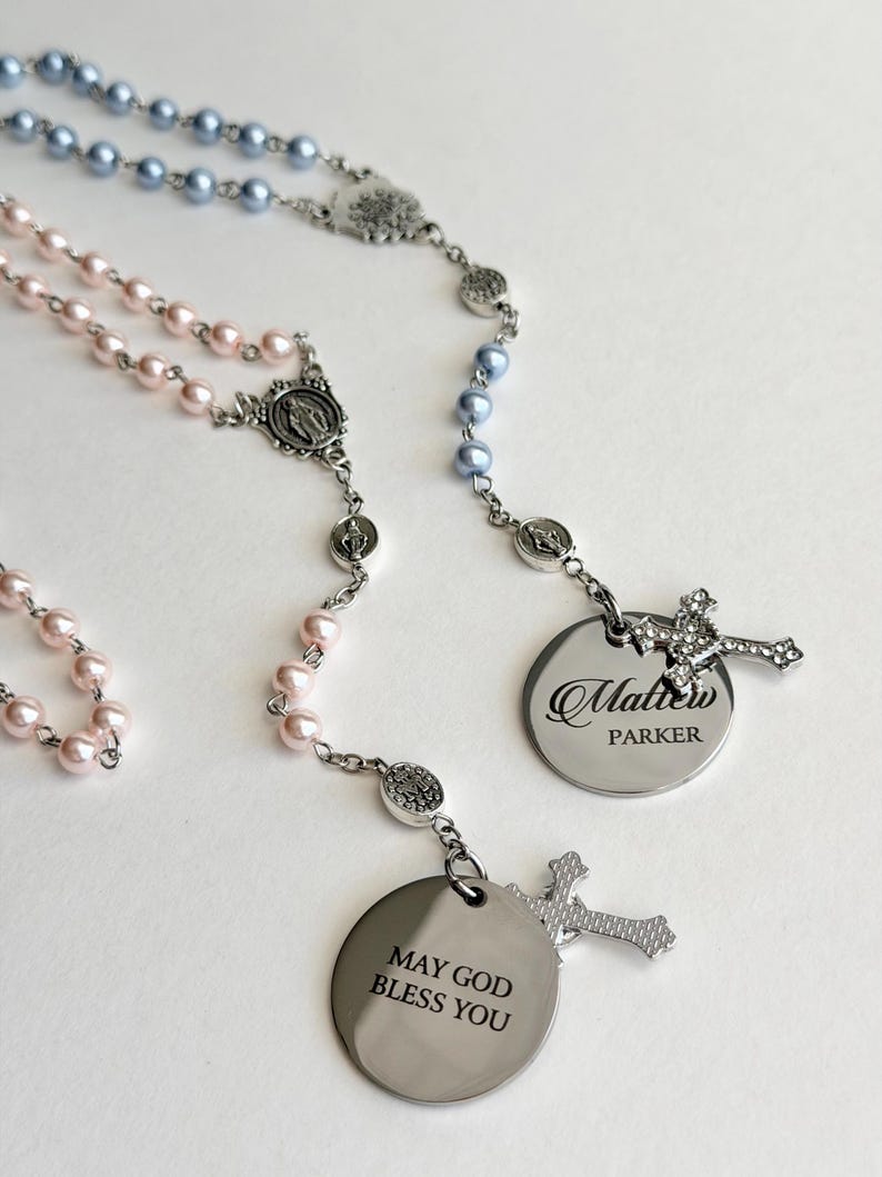 May include: Two rosaries featuring light blue and light pink pearl-like beads on silver-toned chains. Each rosary includes a silver-toned cross, a religious medal, and a round metal pendant. One pendant reads Matthew PARKER, the other MAY GOD BLESS YOU.