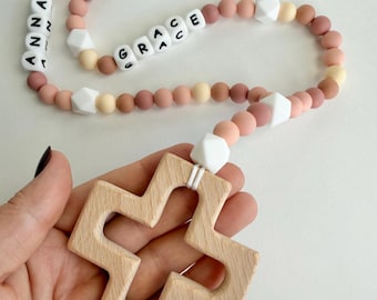 May include: A baby teething necklace with a wooden cross pendant. The necklace features a mix of pink, white, and beige silicone beads, with the names "ANNA" and "GRACE" spelled out in letter beads. The cross is made of light-colored wood.