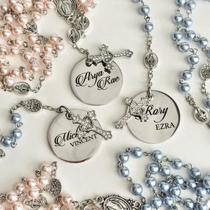 May include: Three rosaries featuring light pink and light blue pearl beads on silver-tone chains. Each rosary includes a silver-tone circular pendant engraved with names: Arya Rae, Rory Ezra, and Mich Vincent, paired with a small crystal-encrusted cross.