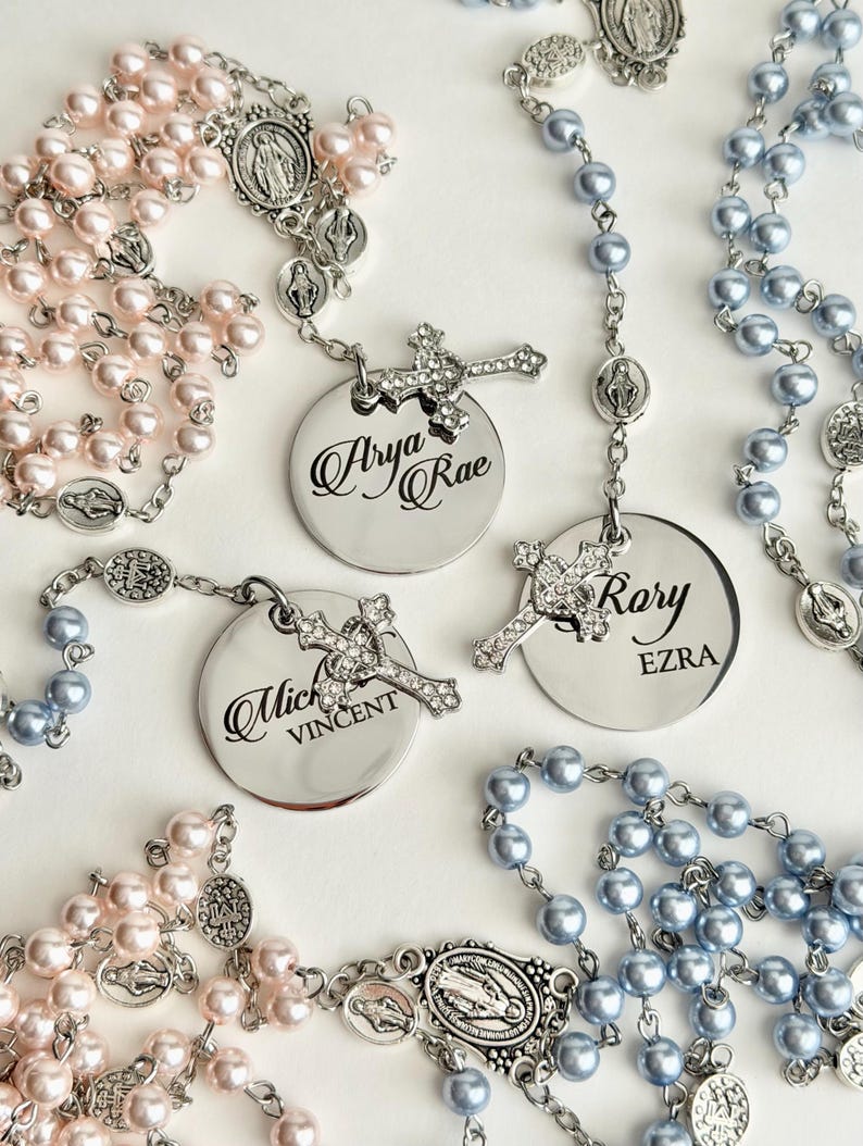 May include: Three rosaries featuring light pink and light blue pearl beads on silver-tone chains. Each rosary includes a silver-tone circular pendant engraved with names: Arya Rae, Rory Ezra, and Mich Vincent, paired with a small crystal-encrusted cross.