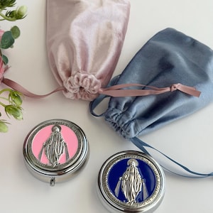 May include: Two round silver-tone metal pill cases featuring a raised figure of the Virgin Mary on a colored enamel background, one pink and one blue. Each case is accompanied by a matching velvet drawstring pouch in soft pink or dusty blue.