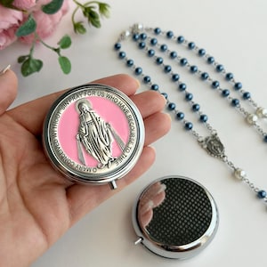 May include: A round silver-toned metal pill box featuring a pink enamel inlay with a raised silver figure of the Virgin Mary and the text O MARY CONCEIVED WITHOUT SIN PRAY FOR US WHO HAVE RECOURSE TO THEE. A second plain silver pill box and a rosary.