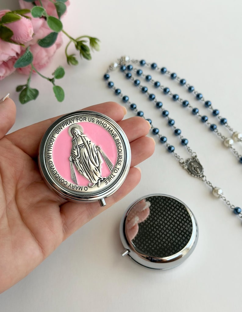 May include: A round silver-toned metal pill box featuring a pink enamel inlay with a raised silver figure of the Virgin Mary and the text O MARY CONCEIVED WITHOUT SIN PRAY FOR US WHO HAVE RECOURSE TO THEE. A second plain silver pill box and a rosary.