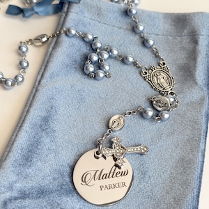 May include: A rosary featuring light blue pearl beads, silver-toned metal links, and religious medals. A silver-toned cross with clear crystals and a round, polished silver-toned metal disc engraved with the name Matthew Parker are attached.
