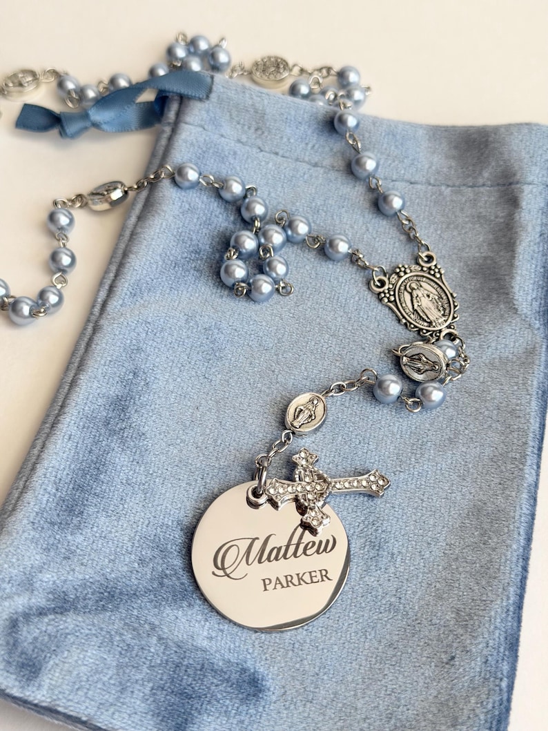 May include: A rosary featuring light blue pearl beads, silver-toned metal links, and religious medals. A silver-toned cross with clear crystals and a round, polished silver-toned metal disc engraved with the name Matthew Parker are attached.