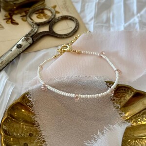 May include: A delicate white beaded bracelet with small pink accents and a gold clasp. The bracelet is displayed on a gold-coloured dish with a pink fabric. Vintage scissors are in the background.