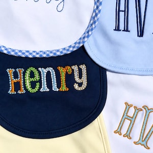 May include: A collection of five baby bibs in white, light blue, navy blue, and pale yellow. The bibs feature embroidered names and initials including Henry and HV in various fonts, colors, and styles. One white bib has a blue gingham trim.