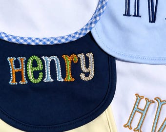 May include: A collection of five baby bibs in white, light blue, navy blue, and pale yellow. The bibs feature embroidered names and initials including Henry and HV in various fonts, colors, and styles. One white bib has a blue gingham trim.