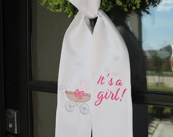 May include: White fabric sash with an embroidered baby carriage and the text "It's a girl!" in pink. The sash is tied to a green wreath, suitable for a baby shower or gender reveal celebration.