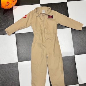 May include: A tan long-sleeve jumpsuit featuring a collar, a front button placket, and long trouser legs. A black rectangular patch with the red text "ROOKIE" is on the chest, and a circular red and white ghost logo patch is on the right sleeve.