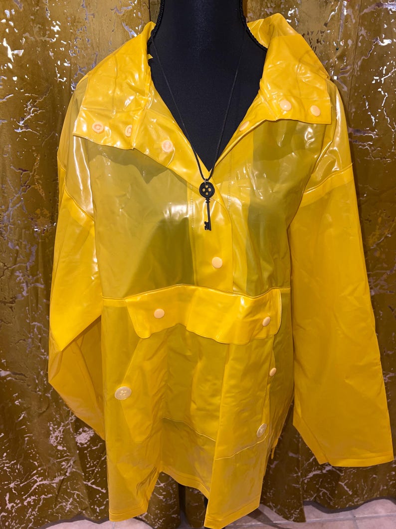K&ouml;nnte beinhalten: Ein leuchtend gelber, transparenter Regenmantel mit Kragen und Knopfverschl&uuml;ssen. Der Regenmantel hat zwei Vordertaschen und eine Schl&uuml;sselanh&auml;nger-Halskette. Das Kleidungsst&uuml;ck wird auf einer schwarzen Schaufensterpuppe pr&auml;sentiert.