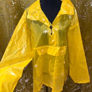K&ouml;nnte beinhalten: Leuchtend gelber, transparenter Regenmantel mit Kapuze und Druckkn&ouml;pfen. Die Jacke hat eine Vordertasche und wird auf einer schwarzen Schaufensterpuppe pr&auml;sentiert. Eine schwarze Schl&uuml;sselanh&auml;nger-Halskette ist sichtbar.
