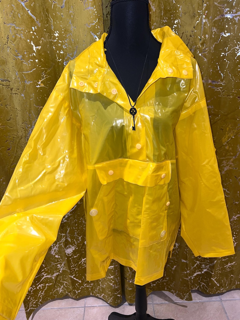 K&ouml;nnte beinhalten: Leuchtend gelber, transparenter Regenmantel mit Kapuze und Druckkn&ouml;pfen. Die Jacke hat eine Vordertasche und wird auf einer schwarzen Schaufensterpuppe pr&auml;sentiert. Eine schwarze Schl&uuml;sselanh&auml;nger-Halskette ist sichtbar.