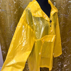 K&ouml;nnte beinhalten: Ein leuchtend gelber, transparenter Regenmantel mit Kragen und Knopfverschl&uuml;ssen. Der Regenmantel hat zwei Vordertaschen und wird auf einer schwarzen Schaufensterpuppe pr&auml;sentiert. Ein schwarzer Schl&uuml;sselanh&auml;nger h&auml;ngt am Hals der Schaufensterpuppe.