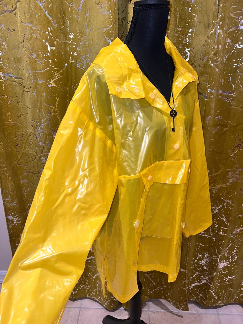 K&ouml;nnte beinhalten: Ein leuchtend gelber, transparenter Regenmantel mit Kragen und Knopfverschl&uuml;ssen. Der Regenmantel hat zwei Vordertaschen und wird auf einer schwarzen Schaufensterpuppe pr&auml;sentiert. Ein schwarzer Schl&uuml;sselanh&auml;nger h&auml;ngt am Hals der Schaufensterpuppe.