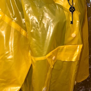 K&ouml;nnte beinhalten: Ein leuchtend gelber Regenmantel mit gl&auml;nzendem Finish. Die Jacke hat einen Kragen, Knopfverschl&uuml;sse und eine Tasche. Ein schwarzer Schl&uuml;sselanh&auml;nger h&auml;ngt am Kragen. Der Regenmantel ist aus wasserdichtem Material.