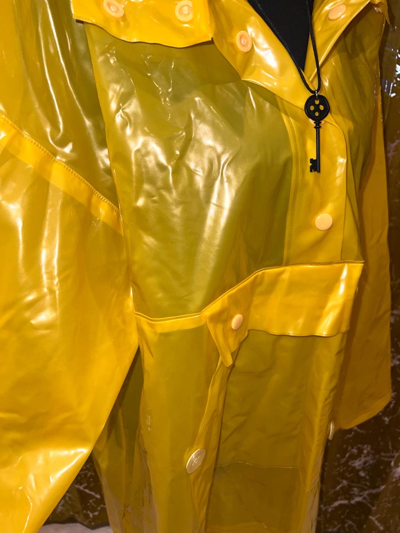 K&ouml;nnte beinhalten: Ein leuchtend gelber Regenmantel mit gl&auml;nzendem Finish. Die Jacke hat einen Kragen, Knopfverschl&uuml;sse und eine Tasche. Ein schwarzer Schl&uuml;sselanh&auml;nger h&auml;ngt am Kragen. Der Regenmantel ist aus wasserdichtem Material.
