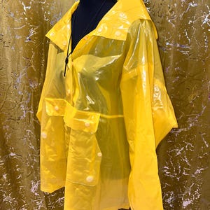 K&ouml;nnte beinhalten: Ein leuchtend gelber, transparenter Regenmantel mit Kapuze und Taschen. Der Regenmantel hat eine Knopfleiste vorne und wird auf einer schwarzen Schaufensterpuppe pr&auml;sentiert. Der Regenmantel besteht aus einem gl&auml;nzenden, wasserdichten Material.
