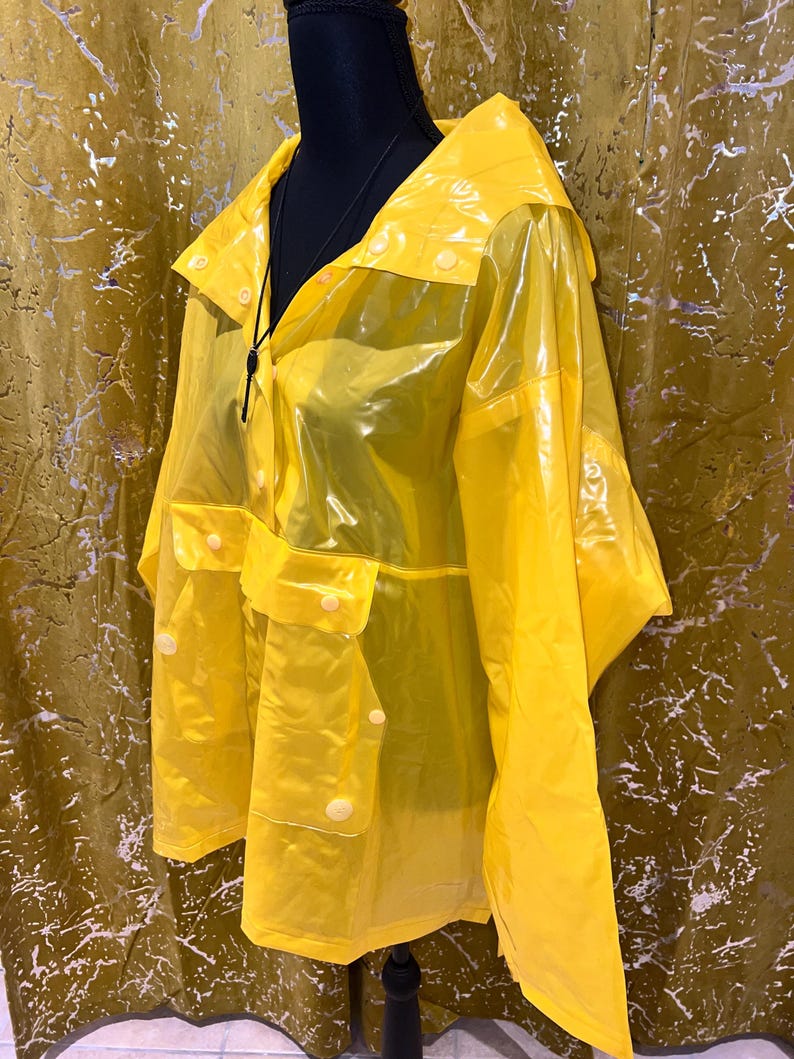 K&ouml;nnte beinhalten: Ein leuchtend gelber, transparenter Regenmantel mit Kapuze und Taschen. Der Regenmantel hat eine Knopfleiste vorne und wird auf einer schwarzen Schaufensterpuppe pr&auml;sentiert. Der Regenmantel besteht aus einem gl&auml;nzenden, wasserdichten Material.