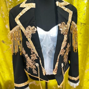 May include: Black blazer with gold trim, floral embroidery, and fringe detailing. The jacket features a white corset-style top underneath. Gold sequins and chains add embellishment to the garment.