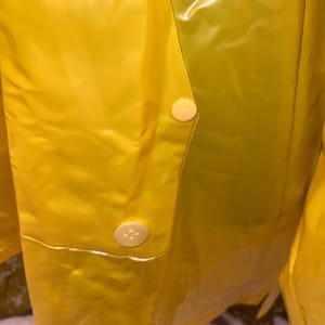 K&ouml;nnte beinhalten: Nahaufnahme eines leuchtend gelben Regenmantels mit zwei cremefarbenen Kn&ouml;pfen. Das gl&auml;nzende, wasserdichte Material reflektiert das Licht und hebt die N&auml;hte und Kn&ouml;pfe hervor. Der Regenmantel scheint im Vintage- oder Retro-Stil zu sein.
