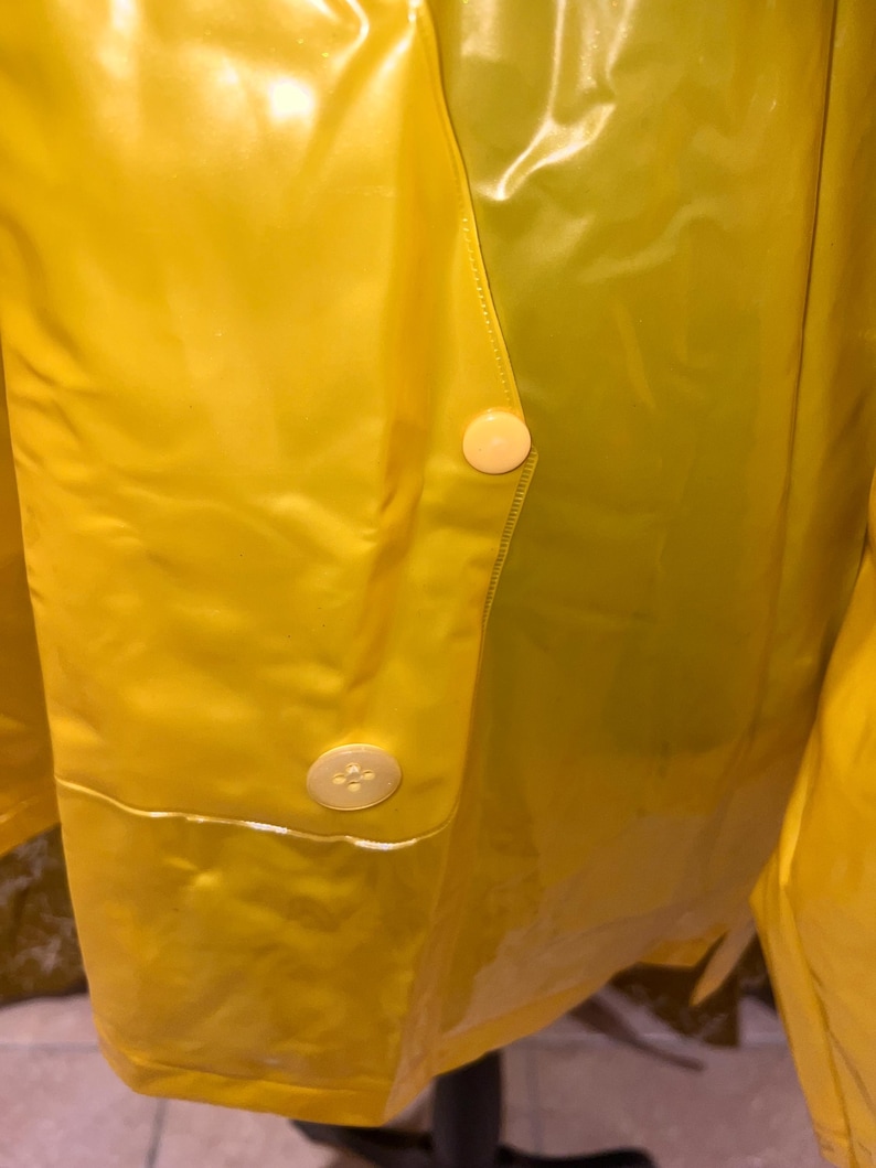 K&ouml;nnte beinhalten: Nahaufnahme eines leuchtend gelben Regenmantels mit zwei cremefarbenen Kn&ouml;pfen. Das gl&auml;nzende, wasserdichte Material reflektiert das Licht und hebt die N&auml;hte und Kn&ouml;pfe hervor. Der Regenmantel scheint im Vintage- oder Retro-Stil zu sein.