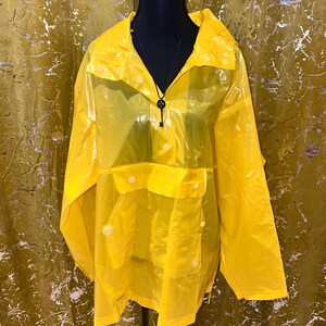 K&ouml;nnte beinhalten: Ein leuchtend gelber, durchscheinender Regenmantel mit Kapuze und Druckkn&ouml;pfen. Der Regenmantel hat eine Vordertasche und wird auf einer schwarzen Schaufensterpuppe pr&auml;sentiert. Eine schwarze Halskette mit einem Schl&uuml;sselanh&auml;nger ist sichtbar.