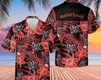 Peut inclure: Une chemise à manches courtes avec un fond noir, un motif floral et de feuilles rouges, et des graphiques de crânes. La chemise porte le mot "Motörhead" en rouge au dos. La chemise est présentée sur une terrasse en bois avec une plage et des palmiers en arrière-plan.