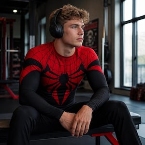 Puede incluir: Una camiseta de compresión de manga larga con un patrón de telaraña rojo y negro en el pecho y los hombros, con un gran emblema de araña negra en el centro. La prenda tiene mangas negras lisas y se combina con pantalones deportivos negros.
