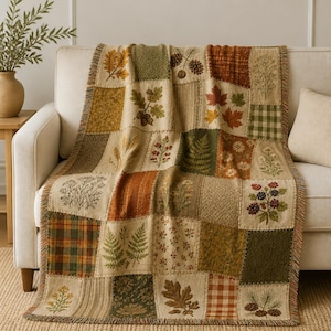 May include: A woven throw blanket featuring a patchwork design with various autumn-themed motifs including oak leaves, acorns, pinecones, ferns, wildflowers, and blackberries in shades of orange, brown, green, and cream with plaid patterns.