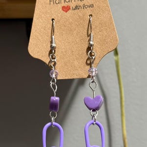 May include: Handmade earrings with a purple safety pin design. The earrings feature a silver chain with purple beads and a heart-shaped charm. The tag reads "Handmade with love."