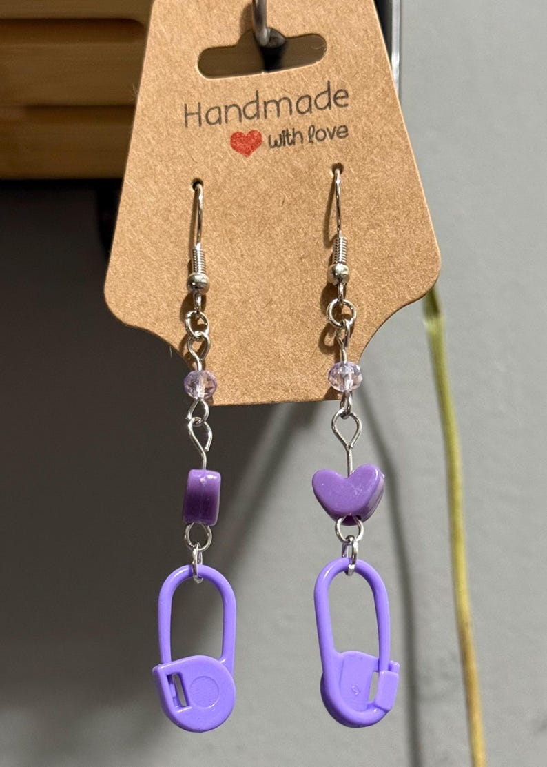 May include: Handmade earrings with a purple safety pin design. The earrings feature a silver chain with purple beads and a heart-shaped charm. The tag reads "Handmade with love."