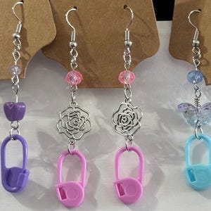 May include: A collection of handmade earrings with silver-tone hooks. The earrings feature various designs, including purple safety pins, pink rose charms, and blue butterfly charms. Each earring has colorful beads and a unique charm.