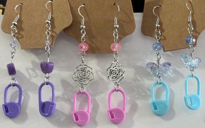 May include: A collection of handmade earrings with silver-tone hooks. The earrings feature various designs, including purple safety pins, pink rose charms, and blue butterfly charms. Each earring has colorful beads and a unique charm.