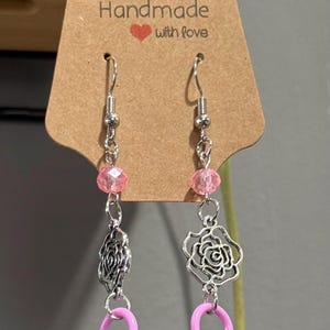 May include: Handmade earrings with a silver chain, pink beads, and a pink safety pin charm. One earring features a silver rose charm, the other a silver filigree charm. The earrings hang from a silver hook. The tag reads "Handmade with love."