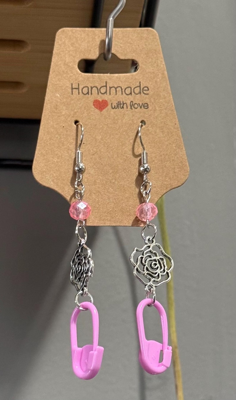 May include: Handmade earrings with a silver chain, pink beads, and a pink safety pin charm. One earring features a silver rose charm, the other a silver filigree charm. The earrings hang from a silver hook. The tag reads "Handmade with love."