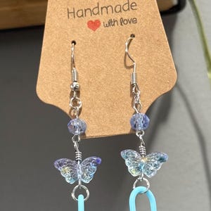 May include: Handmade earrings with a butterfly theme. Each earring features a silver hook, clear beads, a butterfly charm, and a light blue safety pin charm. The tag reads "Handmade with love".