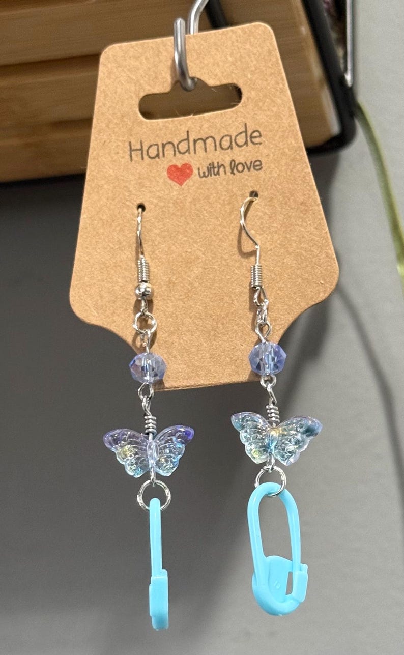 May include: Handmade earrings with a butterfly theme. Each earring features a silver hook, clear beads, a butterfly charm, and a light blue safety pin charm. The tag reads "Handmade with love".