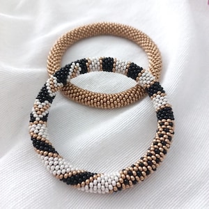 Può includere: Due braccialetti di perline. Uno è di colore oro pieno, e l'altro presenta un motivo di perline nere, bianche e dorate. I braccialetti sono circolari e sembrano fatti a mano, appoggiati su una superficie di tessuto bianco.