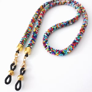 Puede incluir: Una cadena de gafas con cuentas multicolores. La cadena está hecha de pequeñas cuentas de colores y tiene detalles metálicos dorados y bucles de goma negros para sujetar las gafas. La cadena está diseñada para llevarse alrededor del cuello.