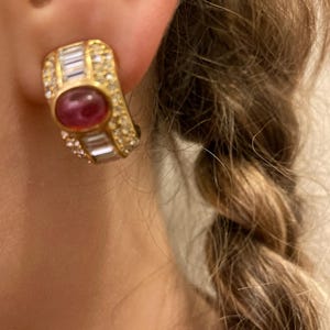 May include: Gold-toned clip-on earring with a central oval ruby and surrounding clear and baguette-cut crystals. The earring is rectangular with rounded edges and is worn on the ear. The design is classic and elegant.