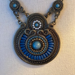 May include: A bronze-toned necklace with a large, circular pendant. The pendant features a central blue stone surrounded by smaller blue and gold beads. The outer ring is detailed with blue rod-shaped beads and decorative metalwork. Two smaller, matching elements are on the chain.