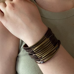May include: A brown leather bracelet with multiple brass-colored metal bars. The bracelet is wrapped around a wrist, showcasing the design and texture of the materials. The bracelet is a fashion accessory.