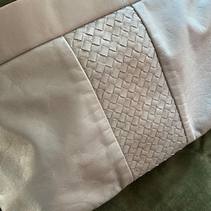 May include: A light beige leather clutch with a woven center panel. The clutch has a rectangular shape with a fold-over top. The leather appears soft and supple, with subtle variations in color and texture.