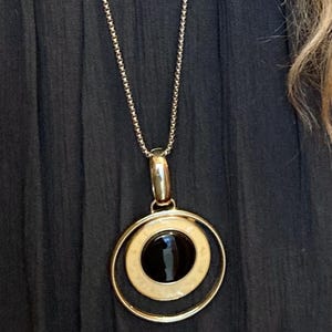 May include: A gold-toned necklace featuring a long, thin chain and a circular pendant. The pendant consists of a large outer gold ring surrounding a smaller, cream-colored circular disc, which frames a smooth, polished black stone in the center.
