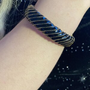 May include: A black bangle bracelet with gold-colored accents. The bracelet has a spiral design with a glossy finish. The gold accents are small, evenly spaced beads that wrap around the bracelet.